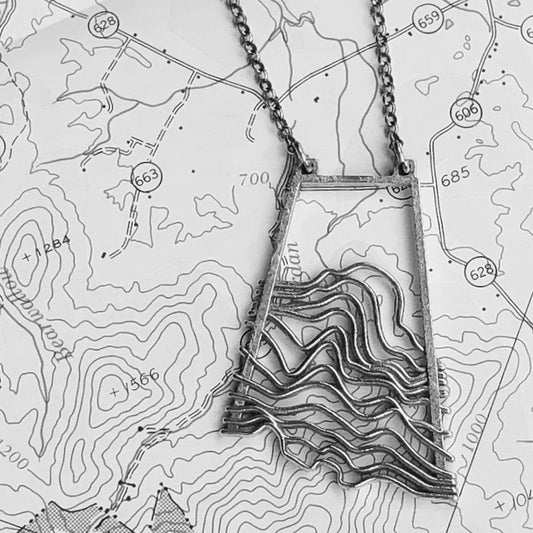 A silver pendant with a geometric wave pattern design,, displayed on a  topographic map.