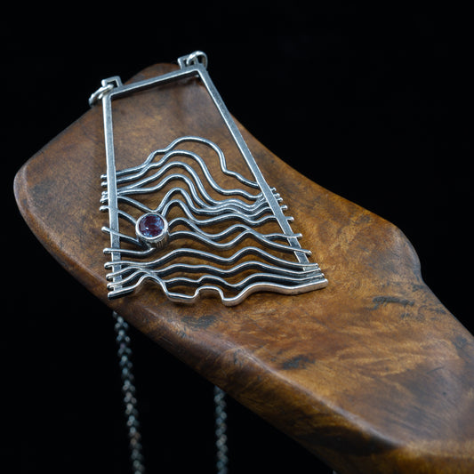 A silver pendant with a geometric wave pattern design, featuring a hand-set gemstone in the center, displayed on a wooden necklace holder.